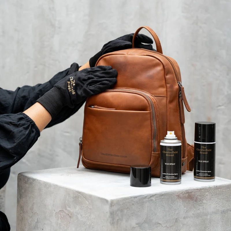 Lederpflege mit The Chesterfield Brand Wax Spray – Hand mit schwarzem Logo-Handschuh pflegt cognacfarbenen Lederrucksack, zwei Sprühflaschen daneben