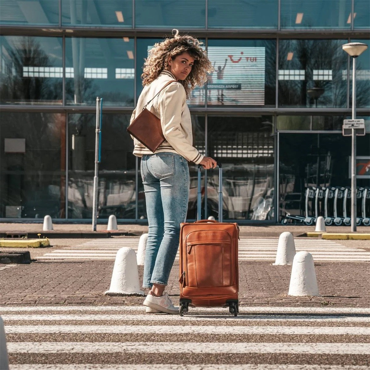 The Chesterfield Brand Trolley-Koffer Key Largo Cognac – Lifestyle-Aufnahme am Flughafen, Frau zieht Leder-Handgepäck über Zebrastreifen