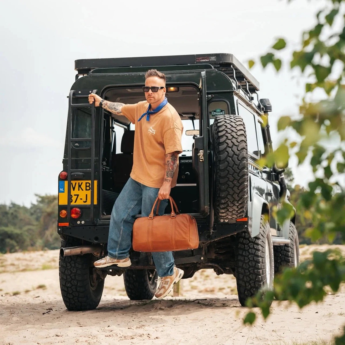 Mann mit The Chesterfield Brand Weekender Perth Cognac – Lifestyle-Aufnahme vor Land Rover Defender in der Natur